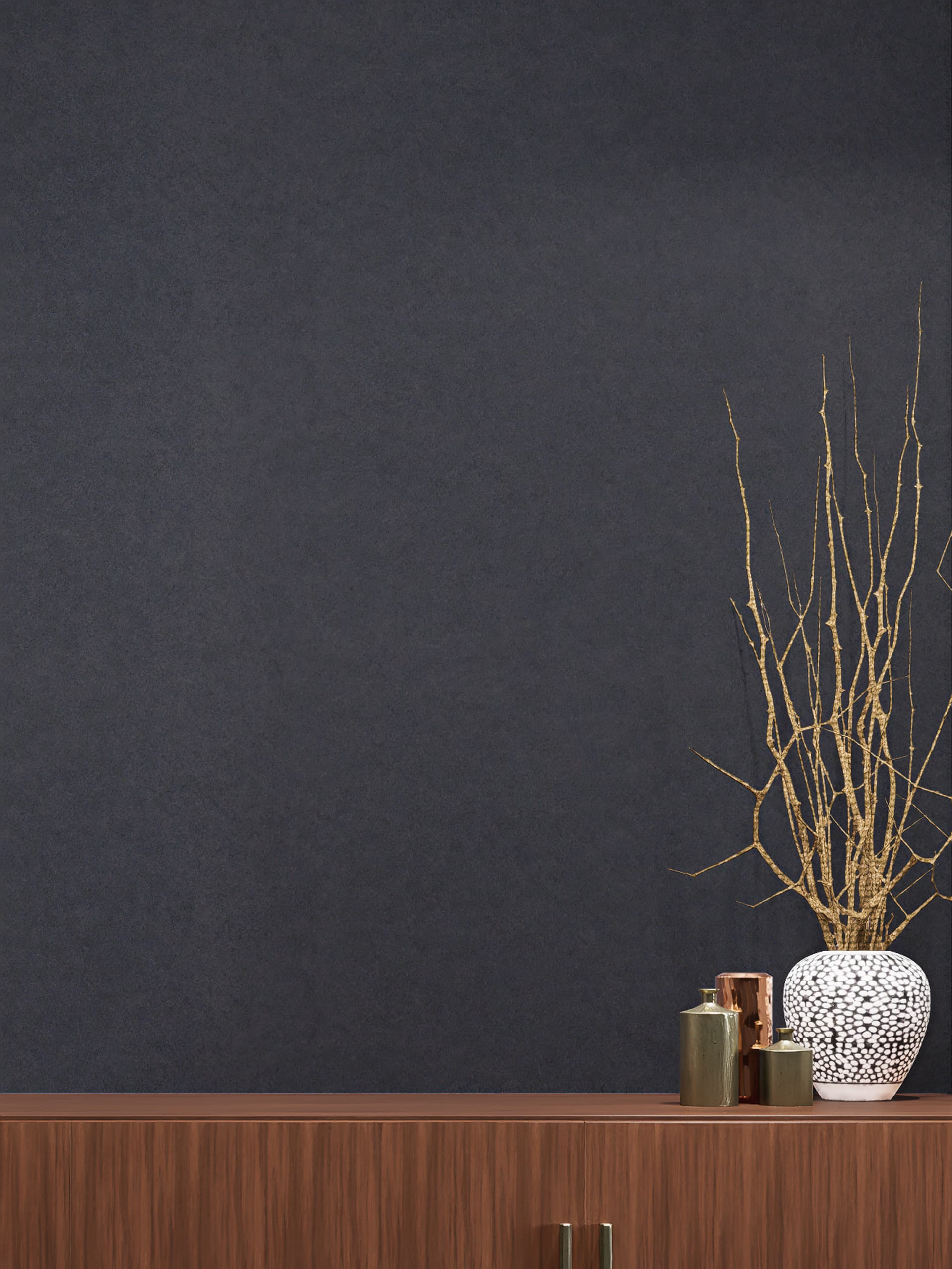 A minimalist scene features a dark grey acoustic felt wall panel behind a wooden cabinet. On the cabinet, there is a white vase with tall, thin branches, accompanied by three small metallic containers in varying sizes. The setting is simple and modern.