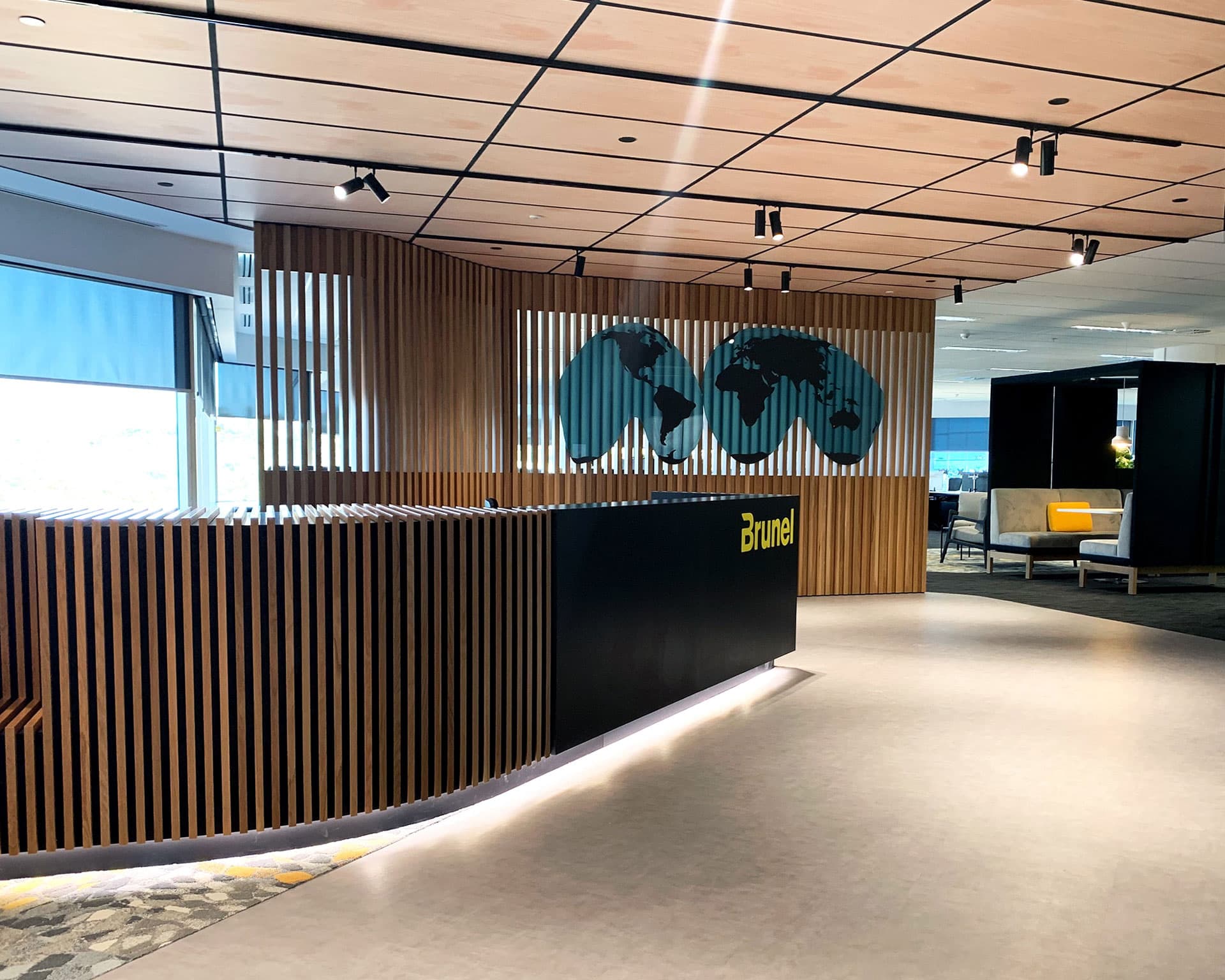 A modern office lobby with a sleek, black reception desk featuring wooden slats and a "Brunel" logo. A stylized world map decorates the wall behind the desk. Natural light streams in through large windows covered by blue blinds, while printed acoustic felt ceiling panels enhance the ambiance. The area is well-lit and clean.