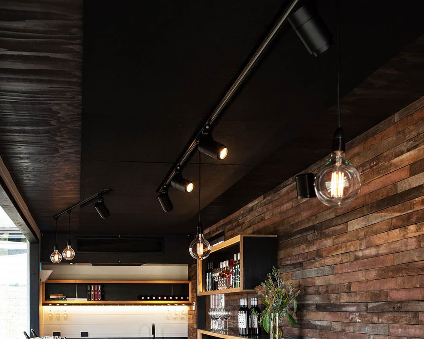 A modern bar interior with wooden panel walls and solid acoustic felt ceiling panels. Exposed light bulbs hang from the ceiling, illuminating the space. Shelves filled with bottles of liquor and wine glasses are visible in the background. A small flower arrangement sits on a shelf.