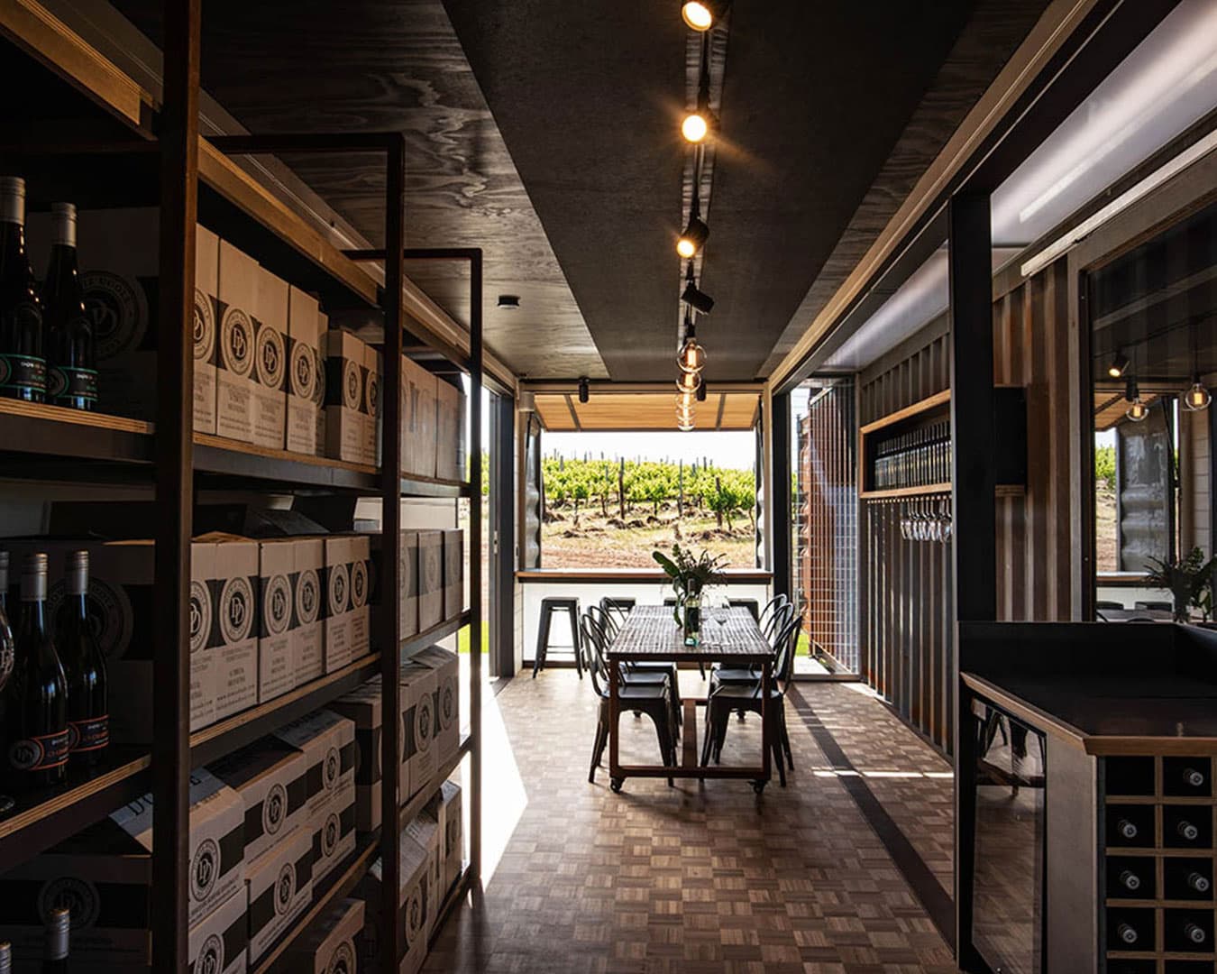 A modern interior of a winery with wooden shelves filled with wine bottles on the left, a long table with chairs in the center, and large windows showing a vineyard outside. The decor includes solid acoustic felt ceiling panels and light bulbs on the ceiling, along with a patterned wooden floor.