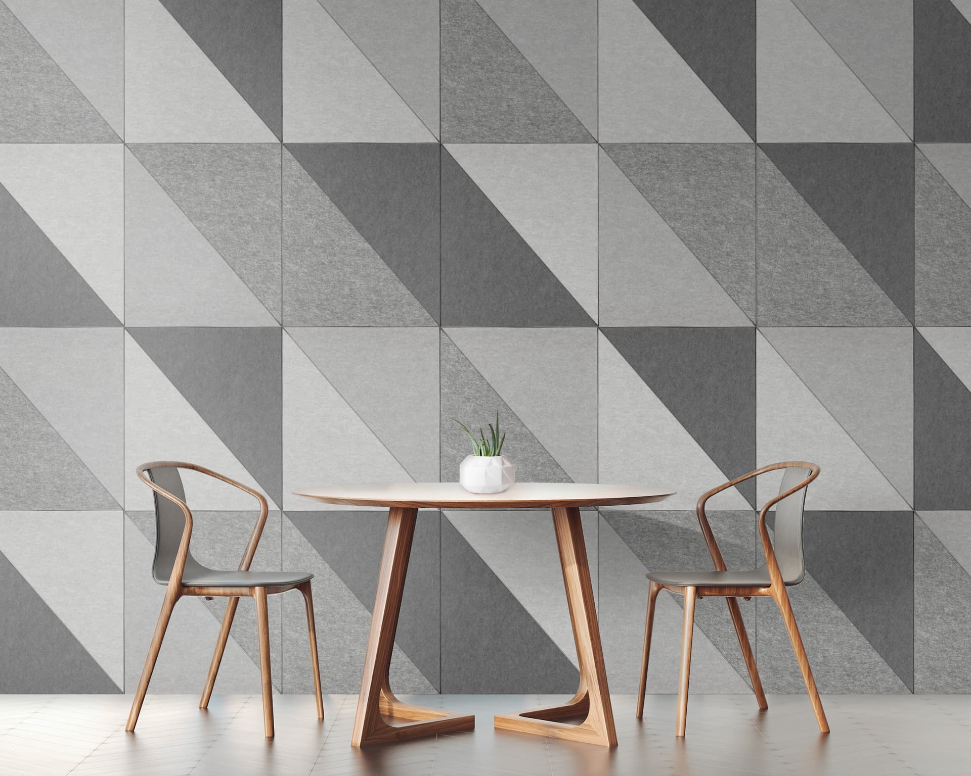 A modern interior featuring a round table with a single white vase holding a small plant. Two wooden chairs with curved backs are positioned on each side. The wall is adorned with acoustic felt wall tiles, arranged in an eye-catching geometric pattern of gray and white triangles.