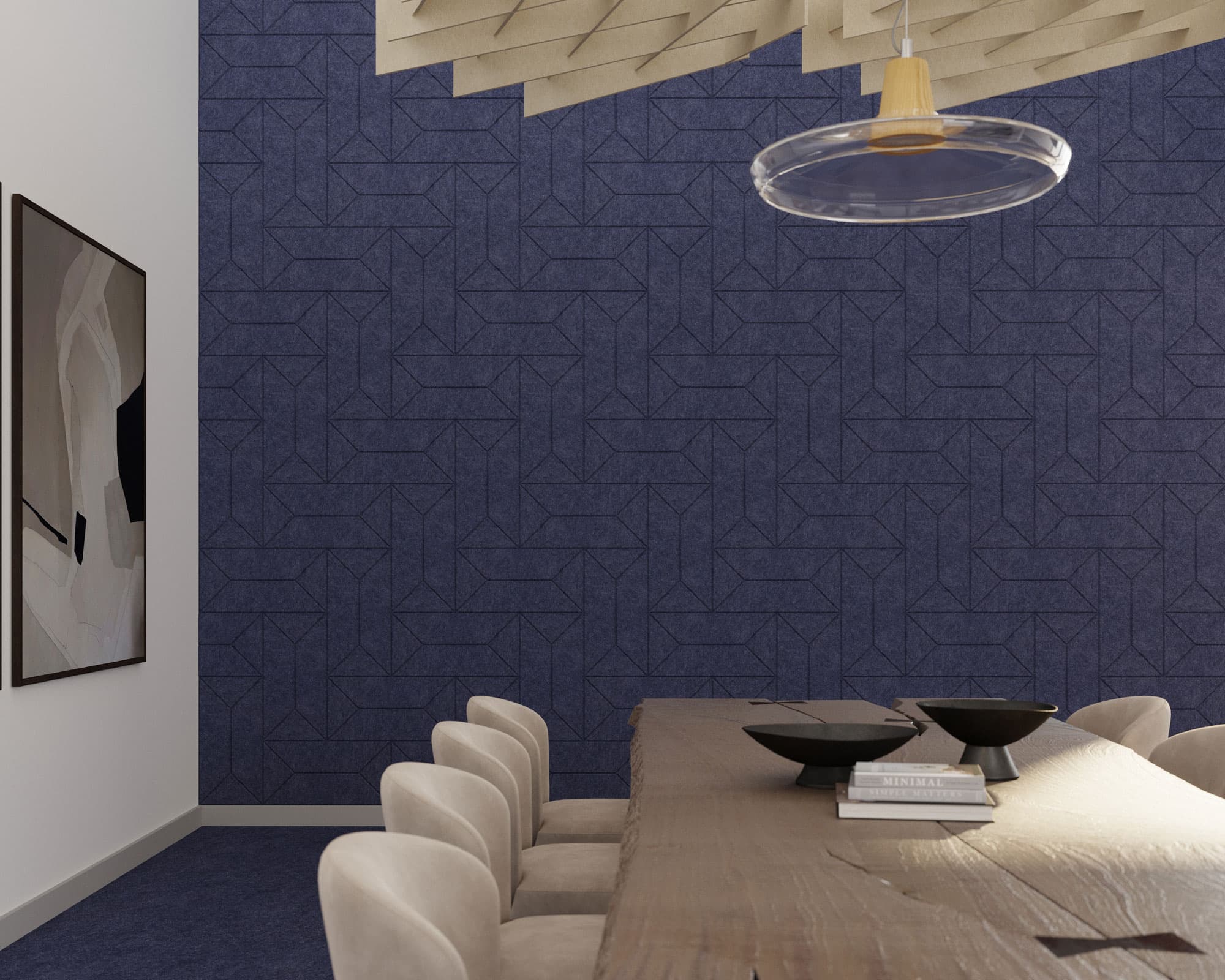 A modern dining room with a wooden table, beige chairs, and a blue geometric patterned accent wall featuring acoustic felt wall tiles. A large abstract painting hangs on a white wall, and a round pendant light with a glass shade hangs above the table. Decorative bowls are placed on the table.