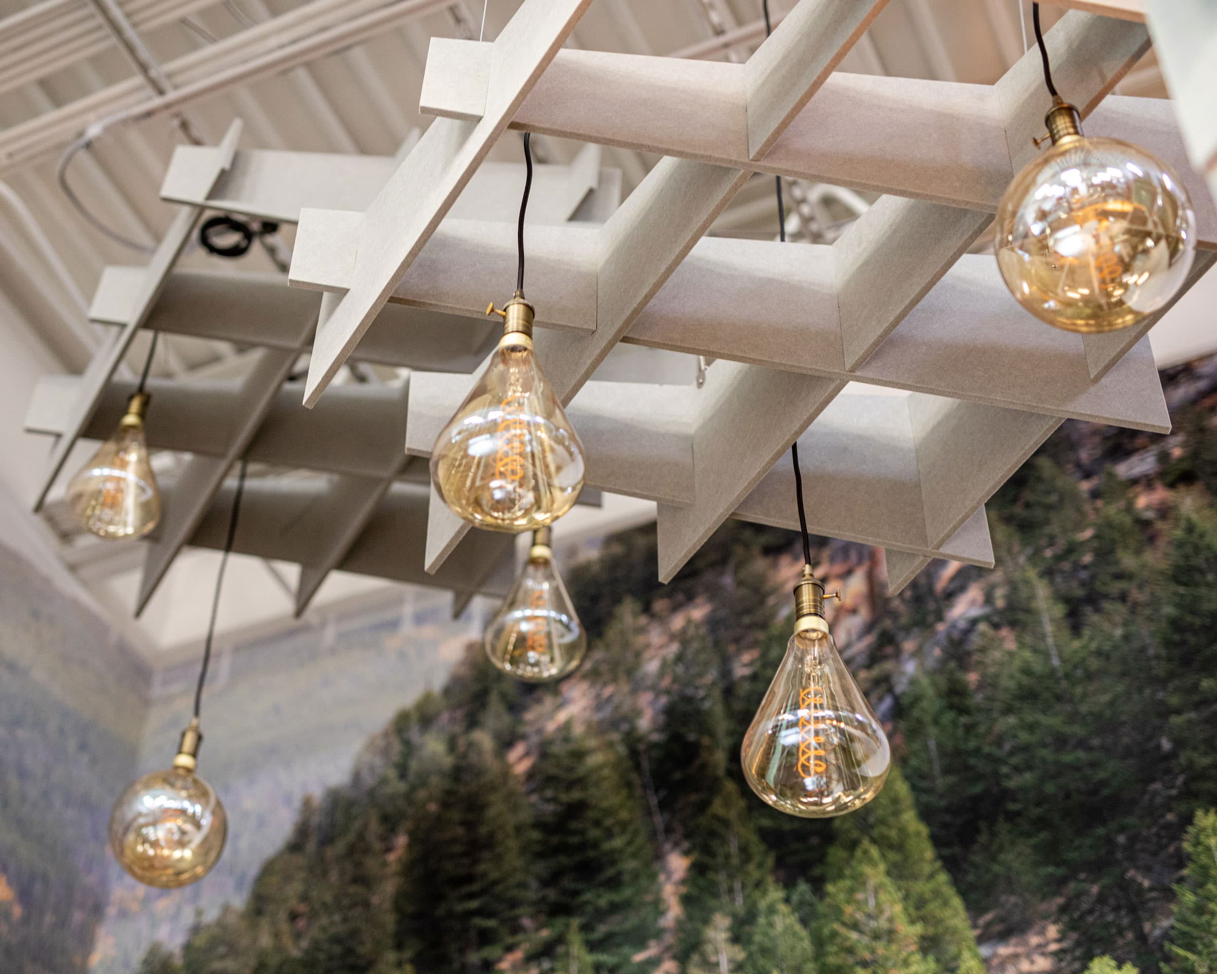 Hanging light bulbs with visible filaments are suspended from a white, geometric ceiling structure. Below, acoustic felt ceiling clouds enhance the sound quality. A forest mural featuring lush evergreen trees decorates the wall in the background, adding a natural ambiance to the space.