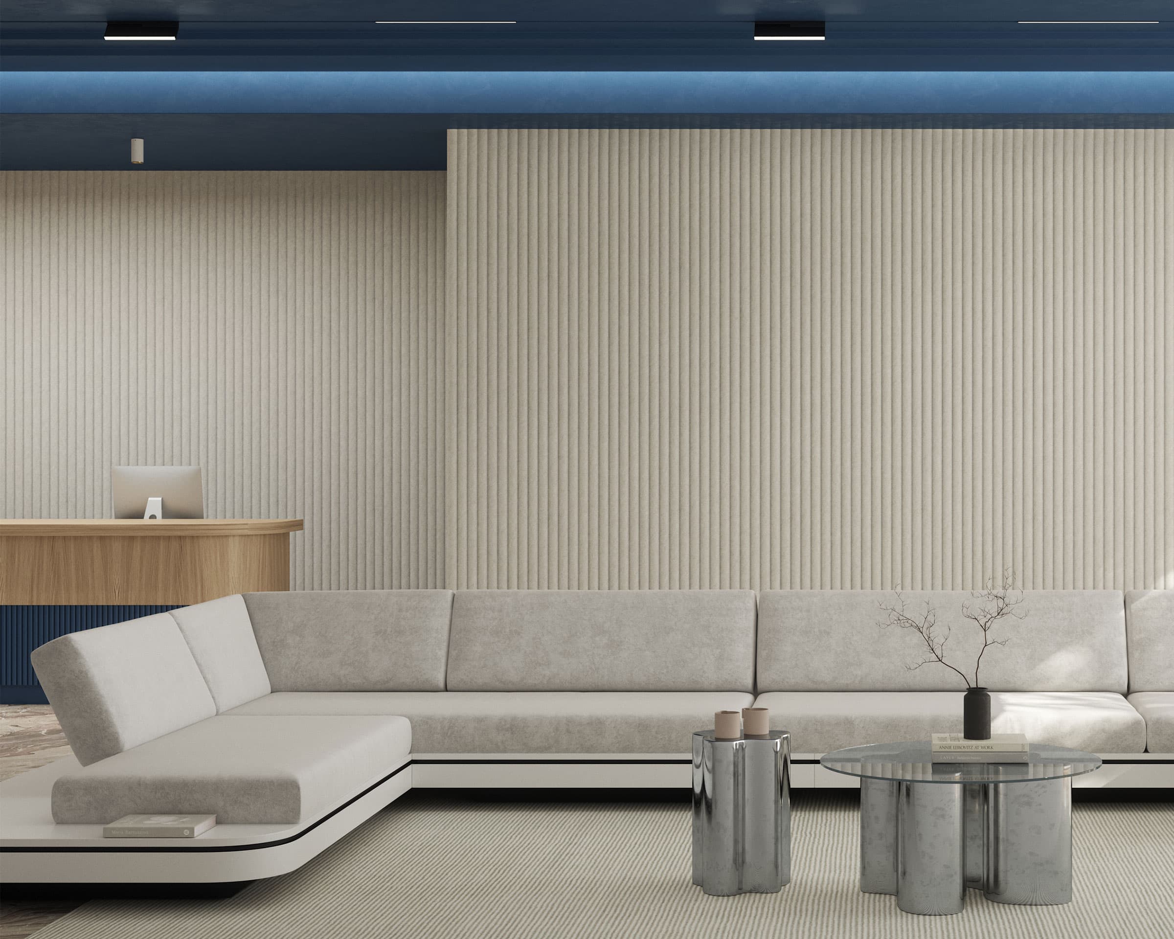 A modern waiting area features a beige sectional sofa with white cushions and a minimalist glass coffee table with silver legs. The backdrop includes celestial acoustic felt wall panels, adding texture to the tall beige wall. A wooden reception desk stands against a blue ceiling in the background.