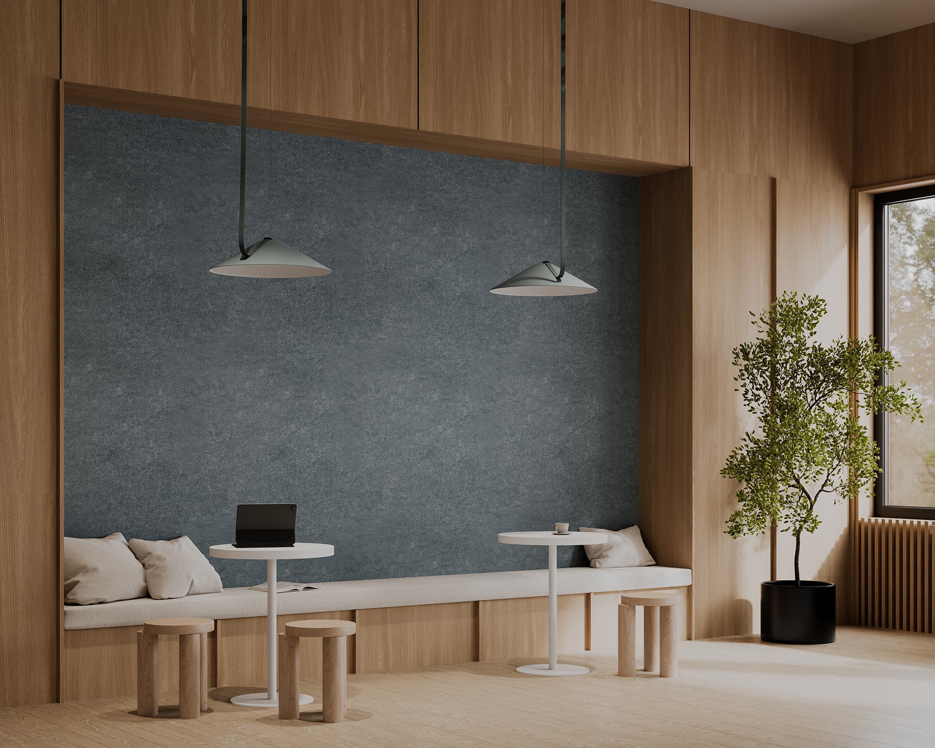 A modern, minimalistic cafe interior featuring a built-in wooden bench with gray cushions and pillows against a grey-green acoustic wall panel. Two round white tables with wooden stools are placed in front of the bench, under two pendant lights. A large potted plant stands by a large window on the right.