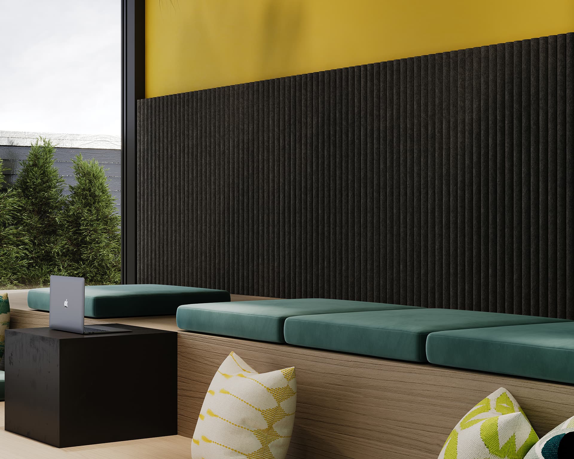 A modern seating area with wooden benches and teal cushion pads is shown. There are yellow and white patterned pillows scattered around. A black side table holds an open laptop. The wall features textured acoustic felt panels on the lower half and yellow paint on the upper half.