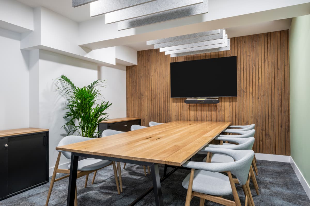 A modern conference room in the GumGum head office boasts a large wooden table surrounded by eight gray chairs. The room features a wood-paneled acoustic felt wall with a flat-screen TV mounted on it. There's a potted plant in the corner and acoustic ceiling panels hanging above the table.