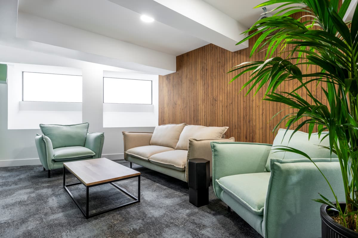 A modern lounge area in the GumGum head office features two light green armchairs, a beige sofa, a wooden coffee table, and a tall potted plant in the corner. The room boasts large windows and a paneled acoustic felt accent wall with gray carpeting on the floor.