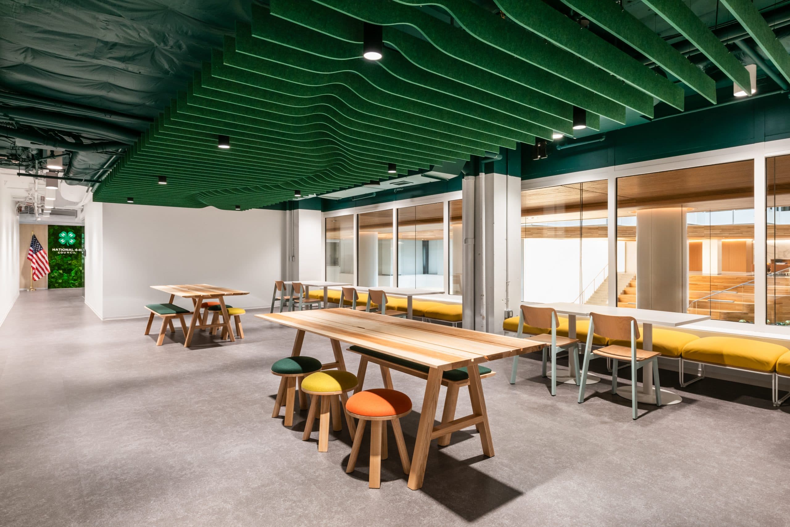 A modern office space with wooden tables and colorful stools features bright lighting that highlights green acoustic ceiling baffles. Spacious glass windows enhance the sleek design, and a visible American flag stands in the corner, adding a touch of pride.