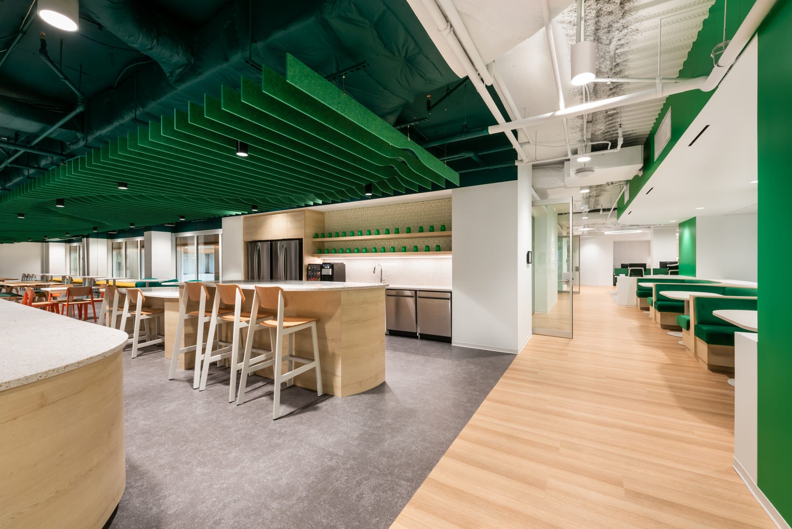Modern office break room with an open layout. Features include a kitchen area with stainless steel appliances, a wooden counter with bar stools, and green acoustic ceiling baffles. Seating booths rest on a wooden floor, while bright lighting enhances the space.