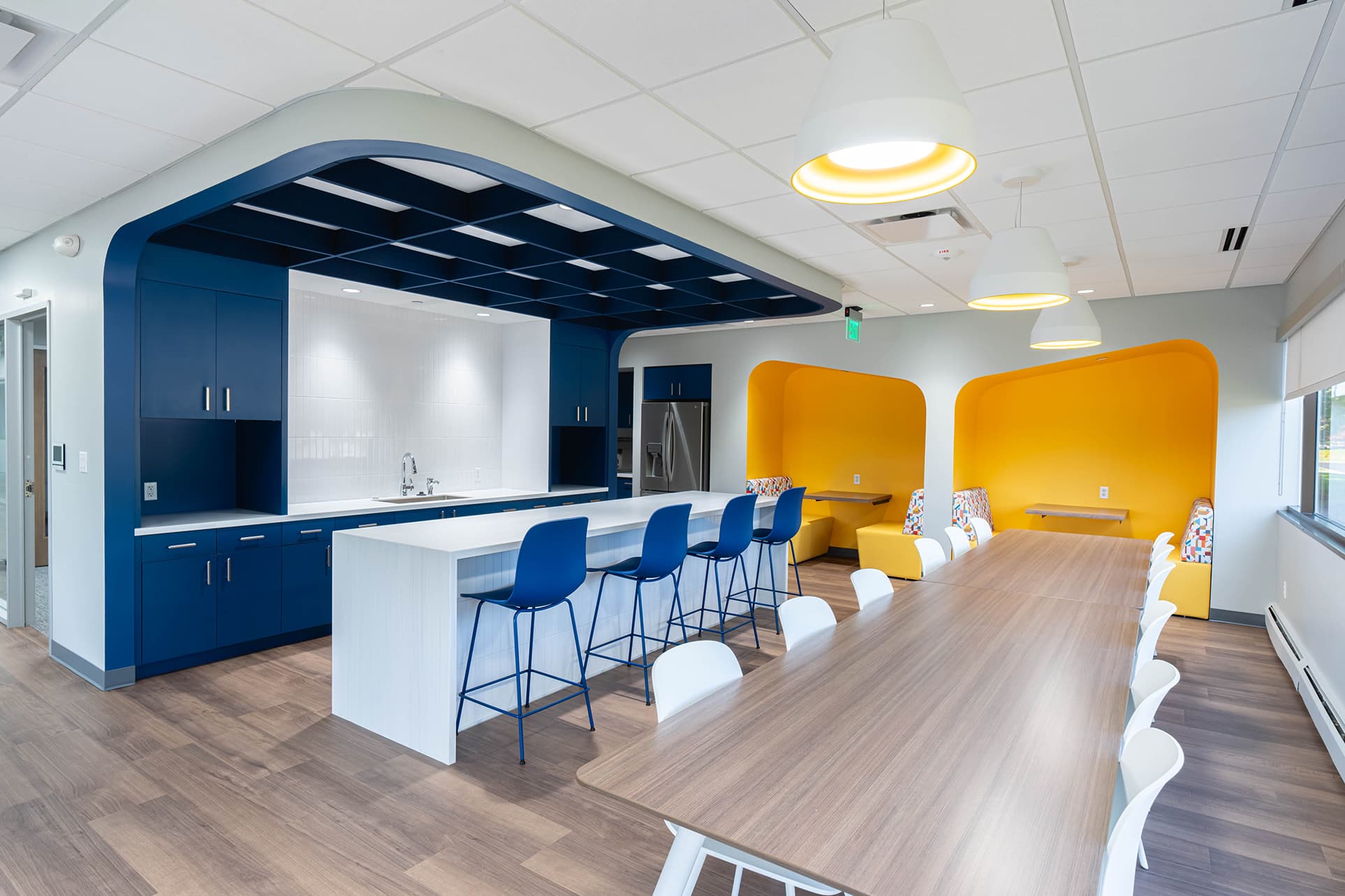 The modern office kitchen and dining area features blue and white cabinetry with an acoustic felt ceiling grid above. A long wooden table with white chairs faces a kitchen island adorned with blue barstools. Yellow alcove seating is bathed in bright overhead lights on the right.