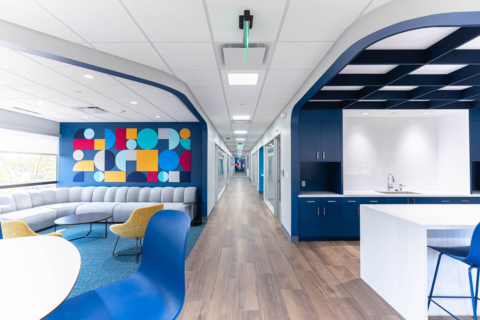 Modern office interior with a long hallway, blue and white color scheme, and acoustic felt grid ceiling. A cozy seating area complements the wooden flooring, which contrasts with the patterned carpet near the kitchenette with stools on the right.