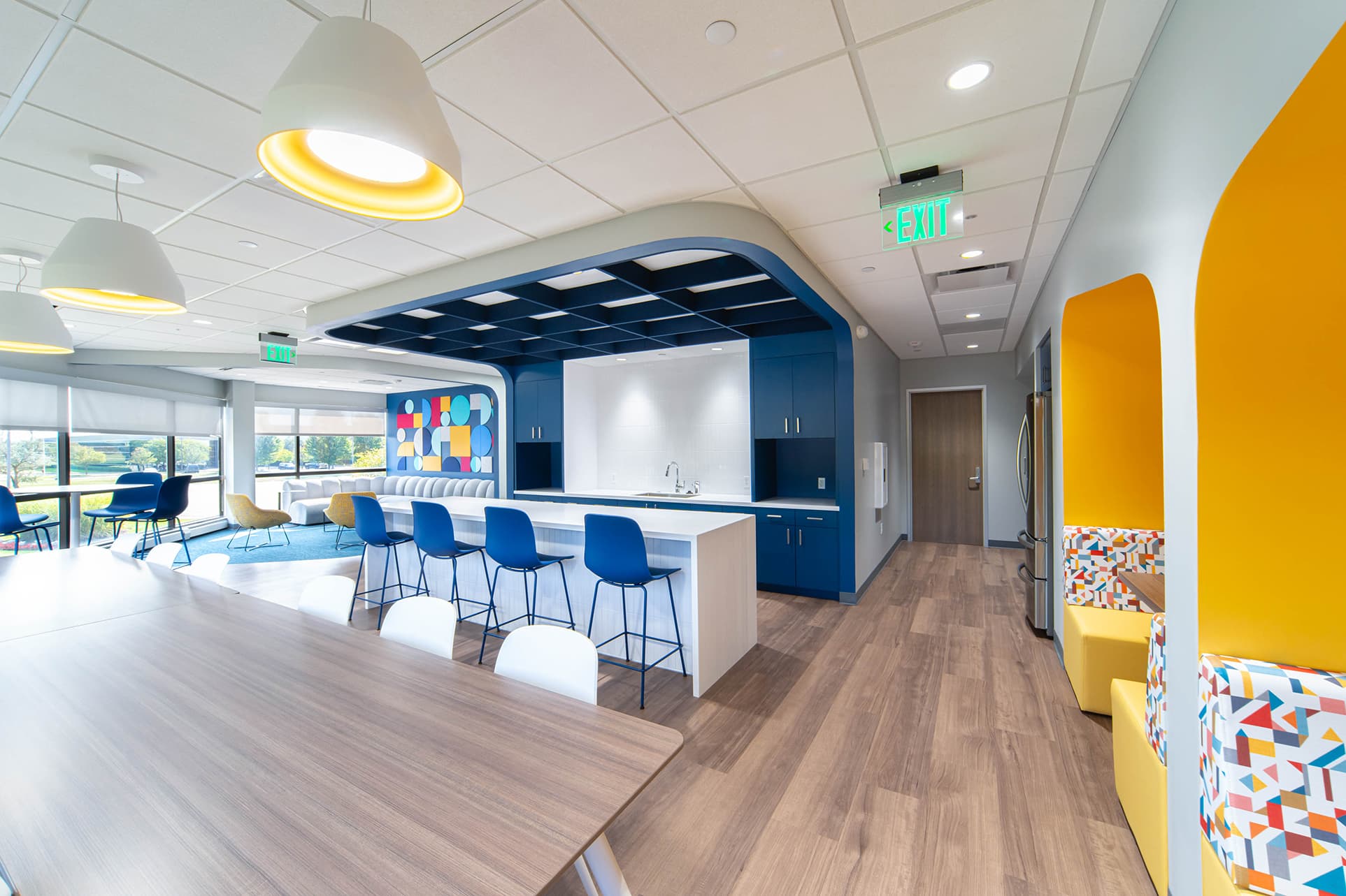 The modern office break room showcases an acoustic felt ceiling grid, and also features wooden flooring and ceiling panels. It includes a kitchen with blue cabinetry, a long counter with blue chairs, colorful geometric art, booth seating with patterned cushions in alcoves, and large windows in the background.