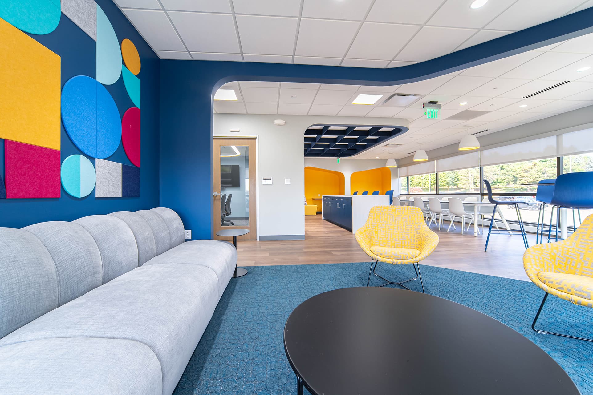 A modern office lounge features a gray sofa, round black table, and yellow chairs under an acoustic felt grid. Colorful geometric wall art adorns the left wall. In the background, a dining area with high tables and chairs enjoys natural light from large windows with chic pendant lights. .