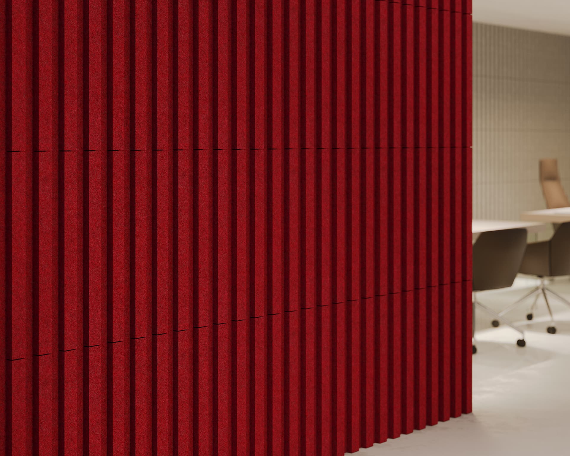 Red vertical panels, made of acoustic felt, adorns a wall, creating a textured design. To the right, a glimpse of an office setting with a table and chairs reveals another wall featuring similar designs in lighter tones, adding a sense of depth and style to the space.