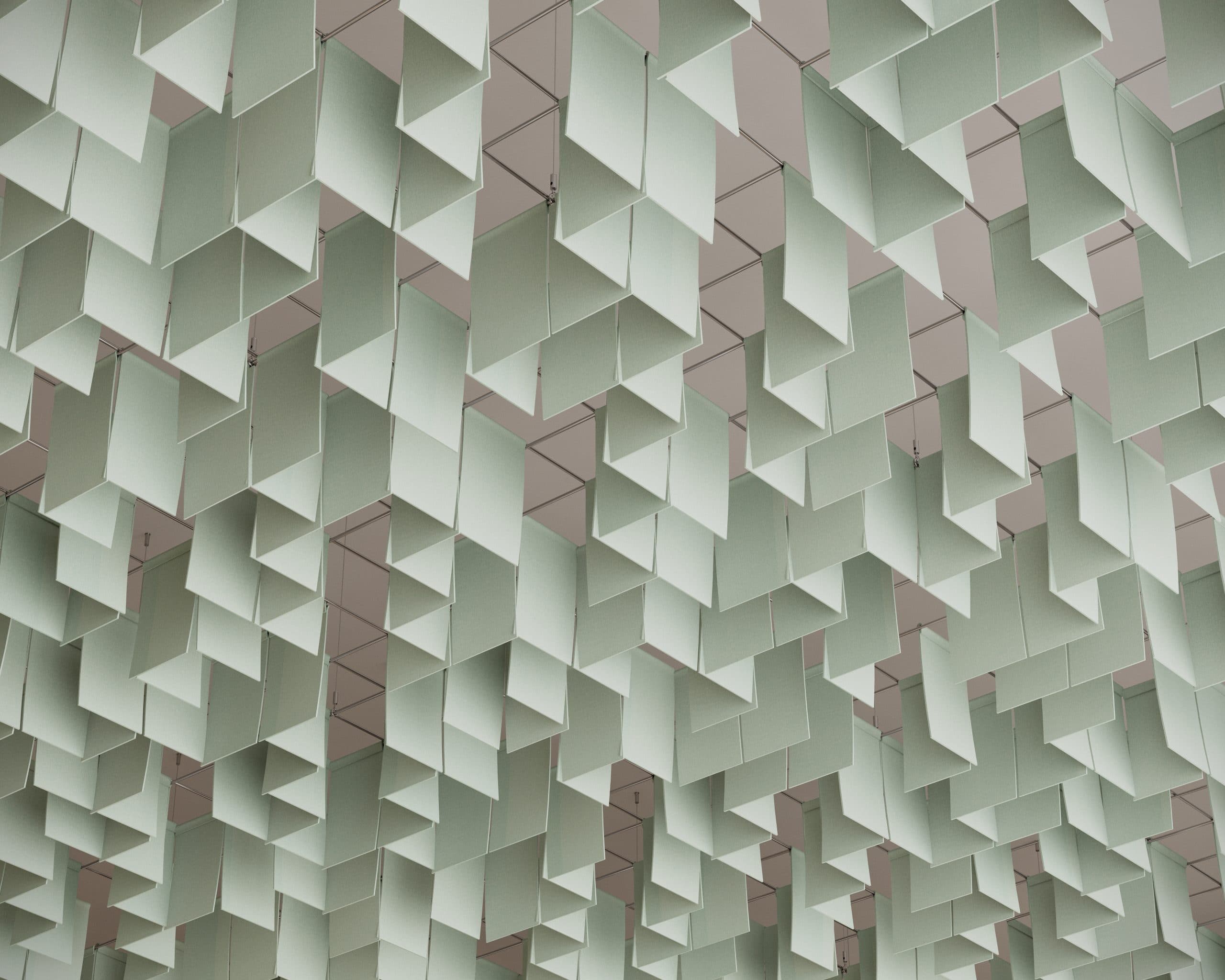 A ceiling art installation features numerous rectangular panels of pale green acoustic felt, suspended at various angles. These panels create a geometric pattern resembling floating clouds, with shadows accentuating the design against the neutral white ceiling backdrop.