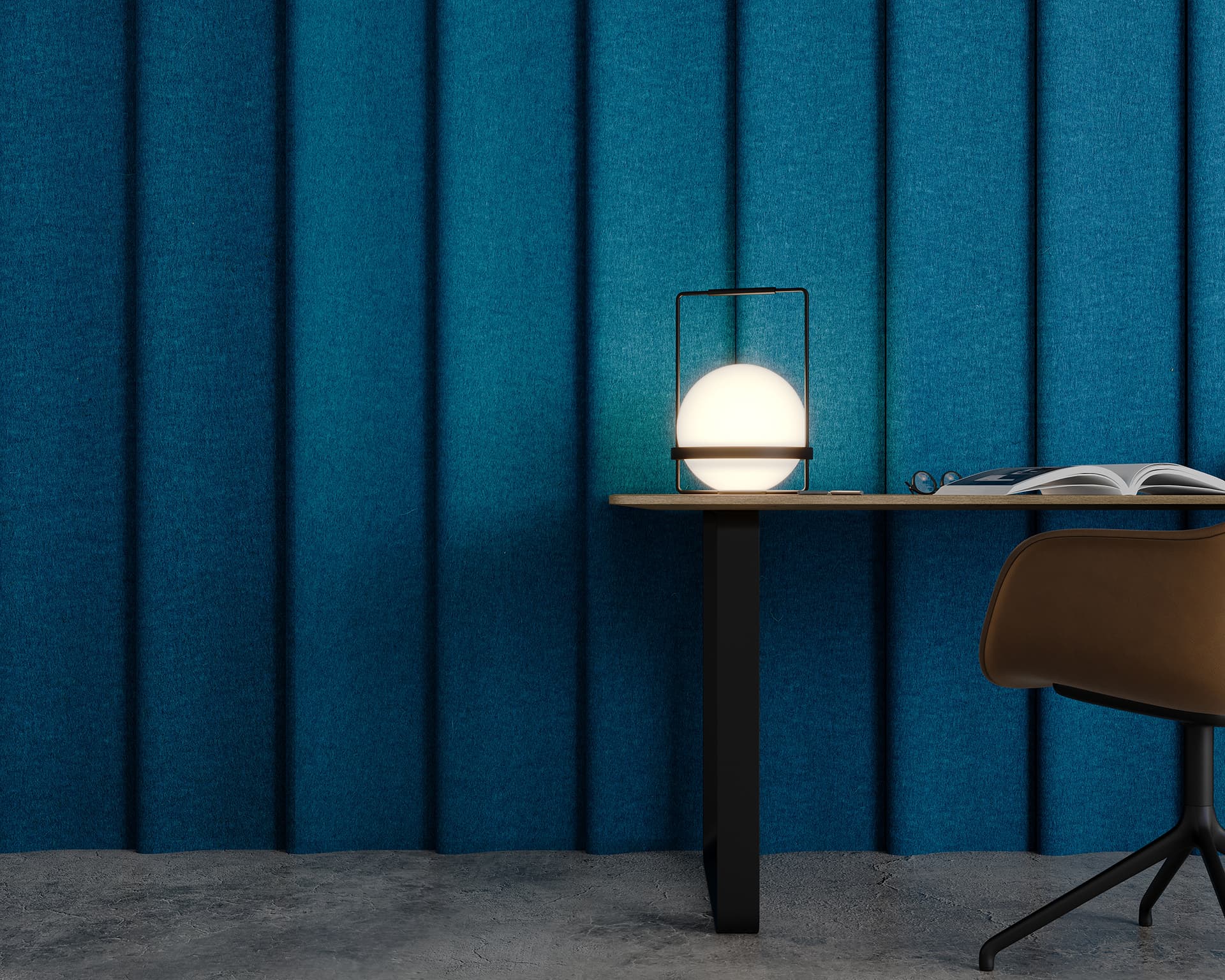 A modern desk setup against a blue textured acoustic felt wall. A spherical lamp with a black frame sits on the desk, casting a warm glow. An open book and glasses are to the right. A brown chair is next to the desk, and the floor is gray concrete.