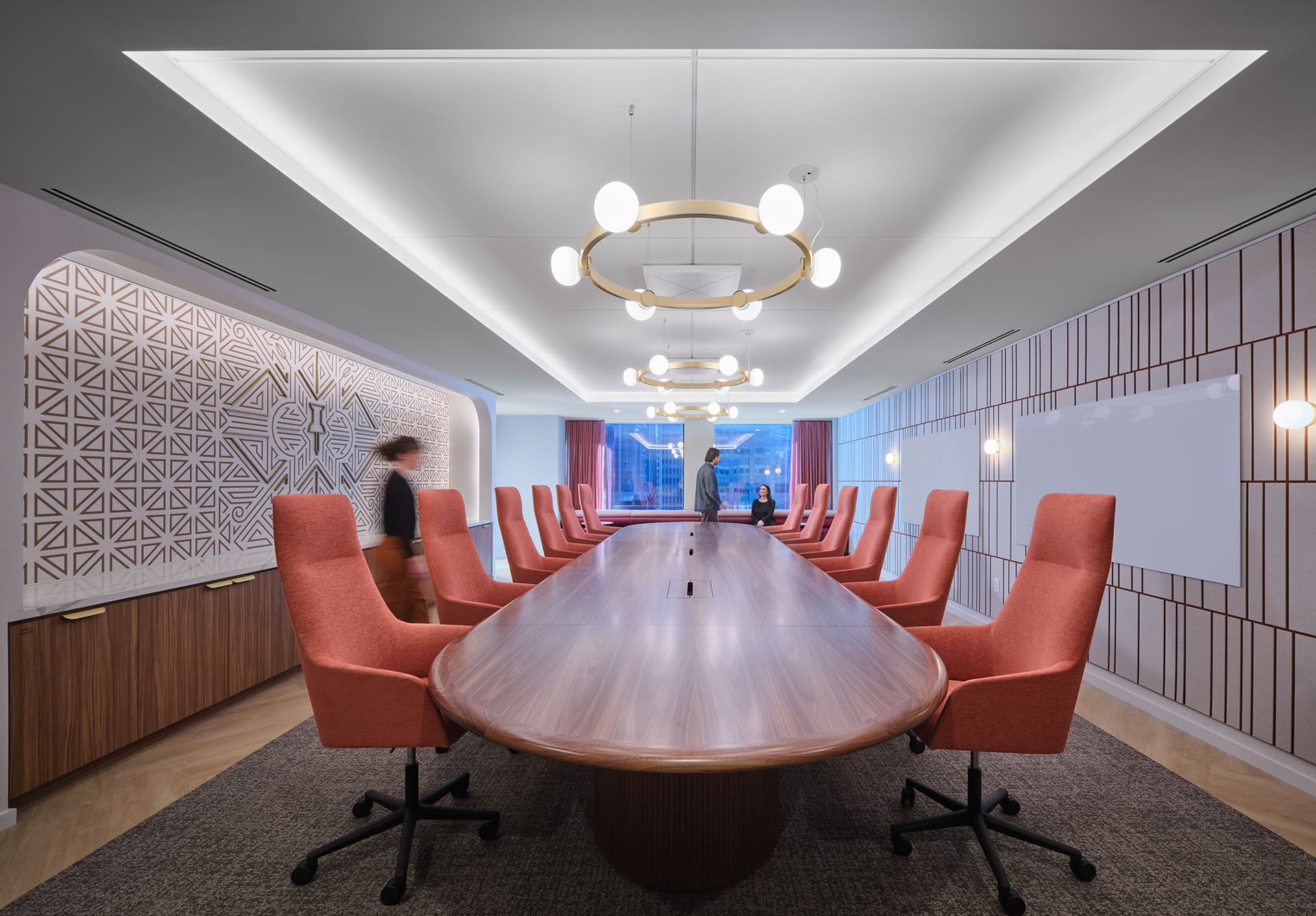 Spacious conference room with a long wooden table, high-back orange chairs, and geometric acoustic felt wall art. Under modern circular light fixtures, two people engage: one by the window, the other standing nearby.