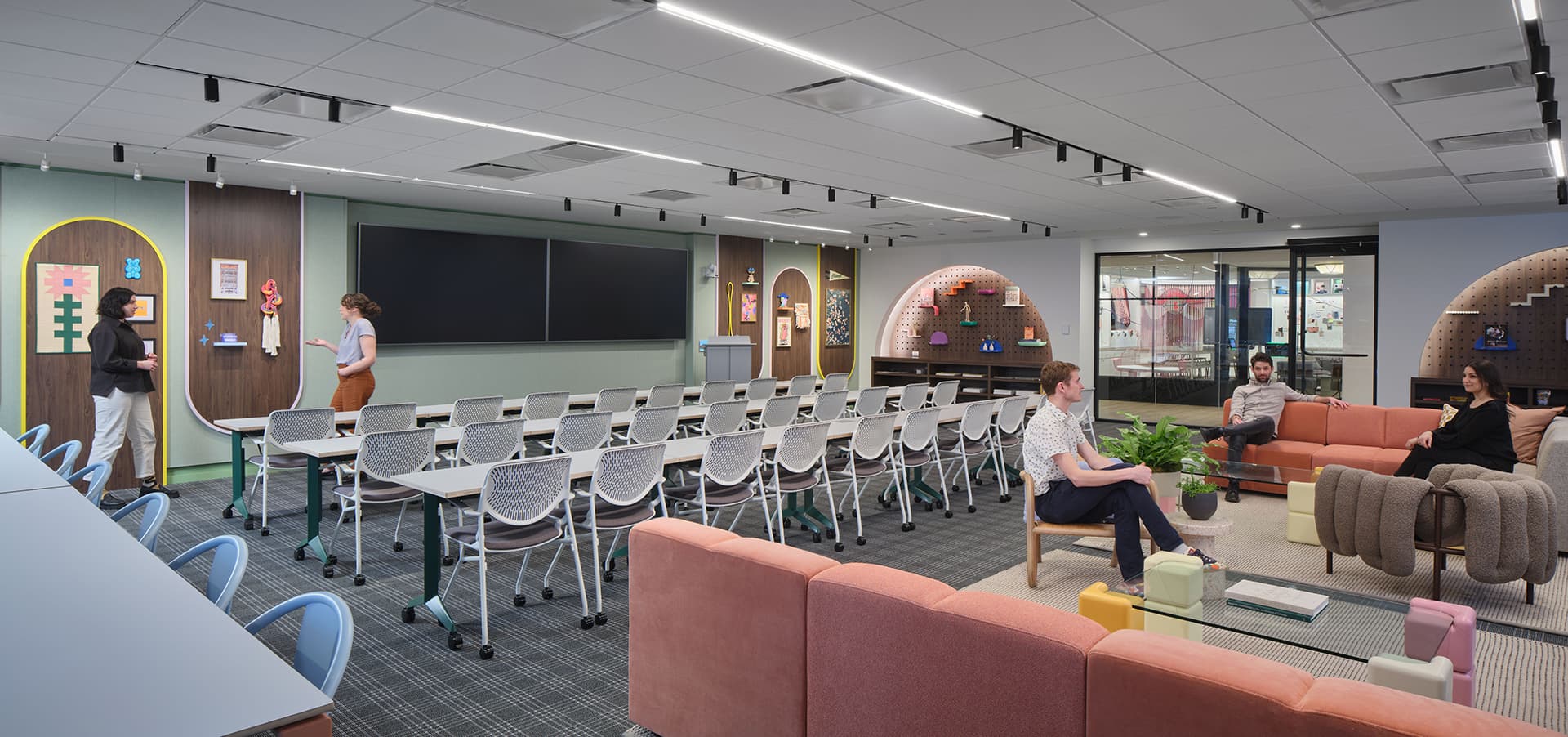 A modern office space with rows of white chairs facing a large screen. People are engaged in conversation. The room features colorful decor, a mix of seating arrangements, and green plants, creating a welcoming atmosphere.