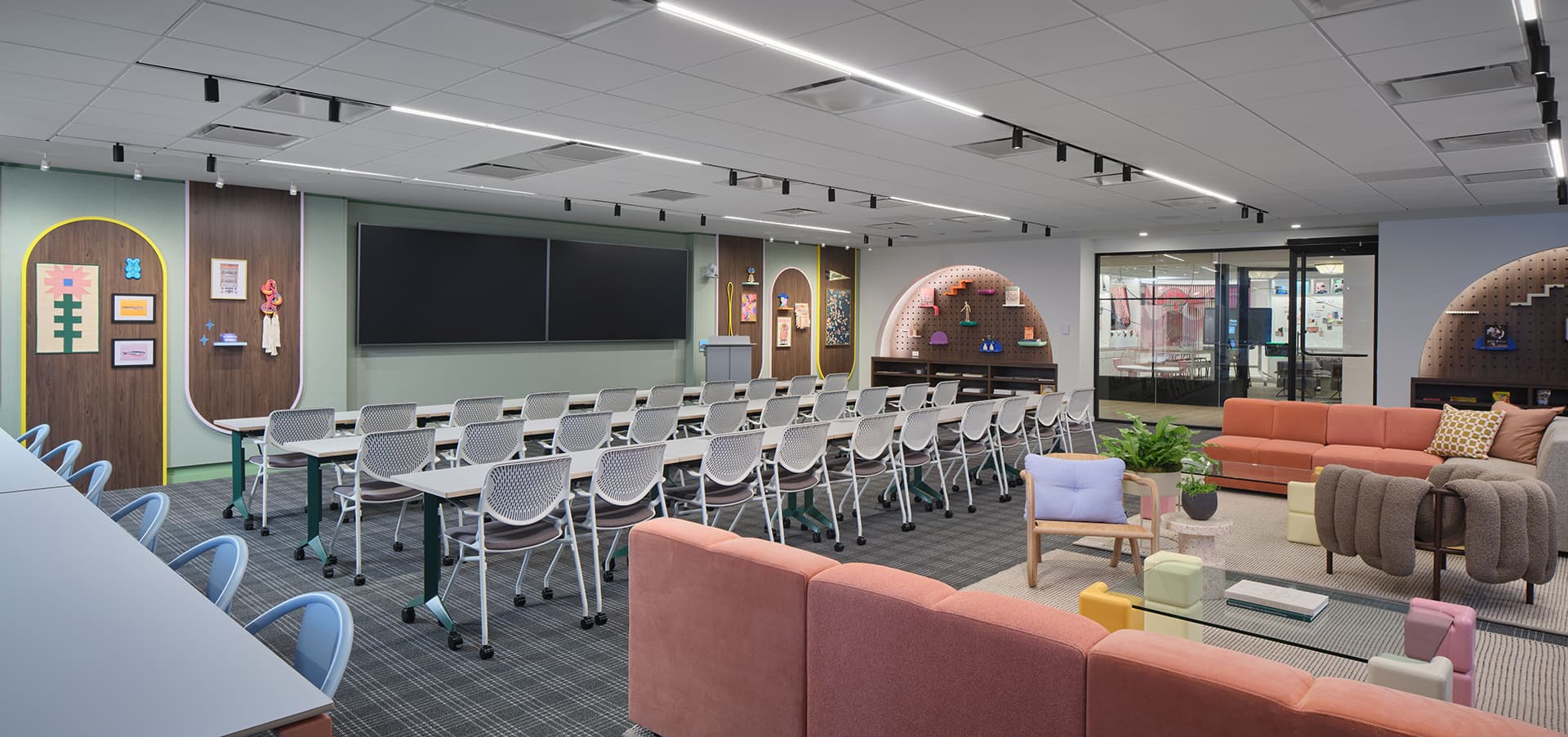 A modern classroom features rows of white desks and chairs facing a large screen. The room boasts colorful, decorative walls, diverse seating areas including couches, and vibrant shelves. Fluorescent lighting adds brightness to this creative space.