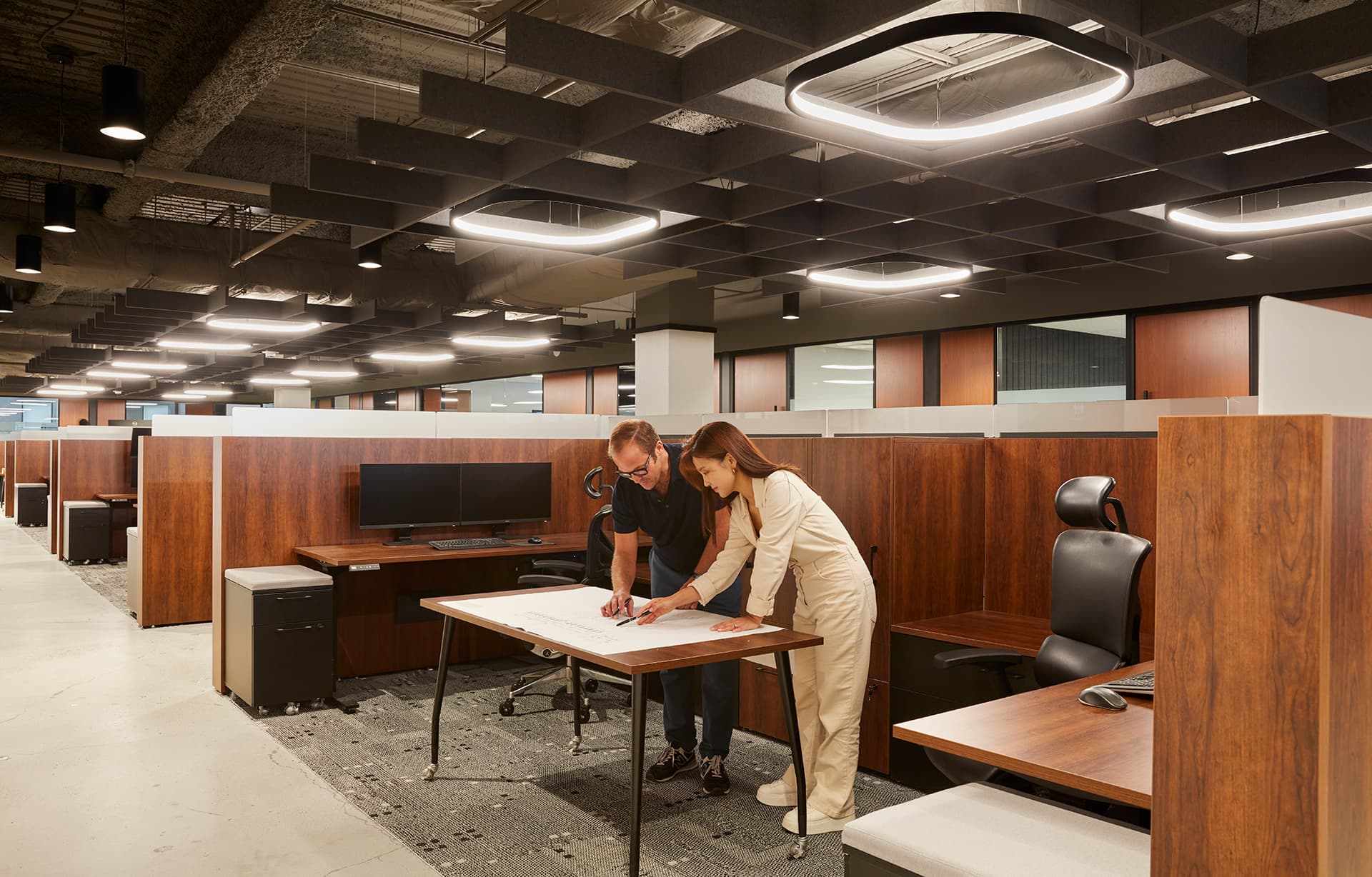 Two people in an office setting, reviewing plans on a table. The office has wooden partitions, a row of desks with computers, and modern circular light fixtures on the ceiling, framed by an acoustic felt ceiling grid. The floor is carpeted with a gray pattern.