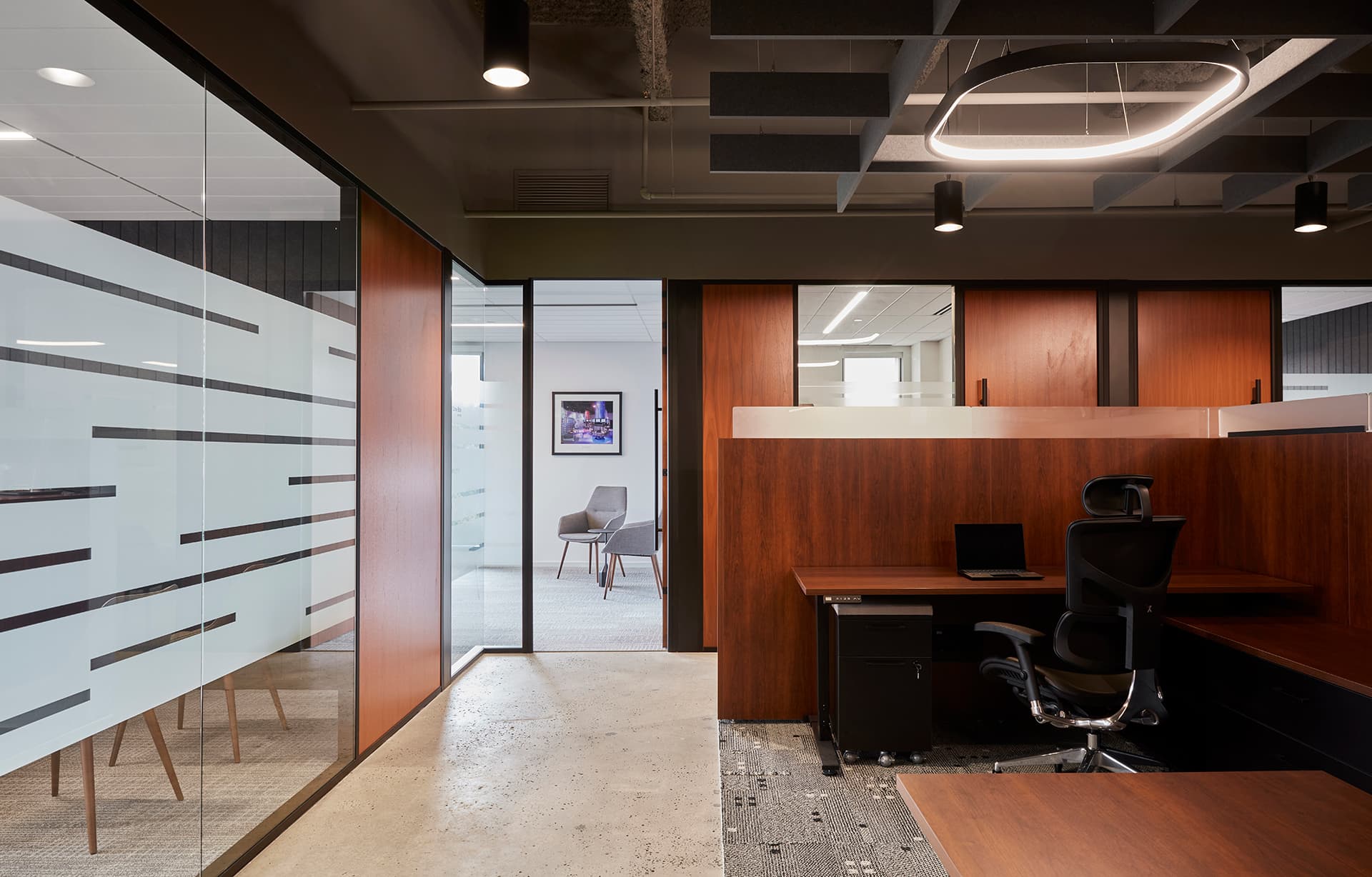 A modern office space featuring wooden desks, a black office chair, and glass partitions with frosted designs. Overhead lighting includes circular and spotlight fixtures within an acoustic felt ceiling grid. A framed picture and chairs are visible in an adjacent room.