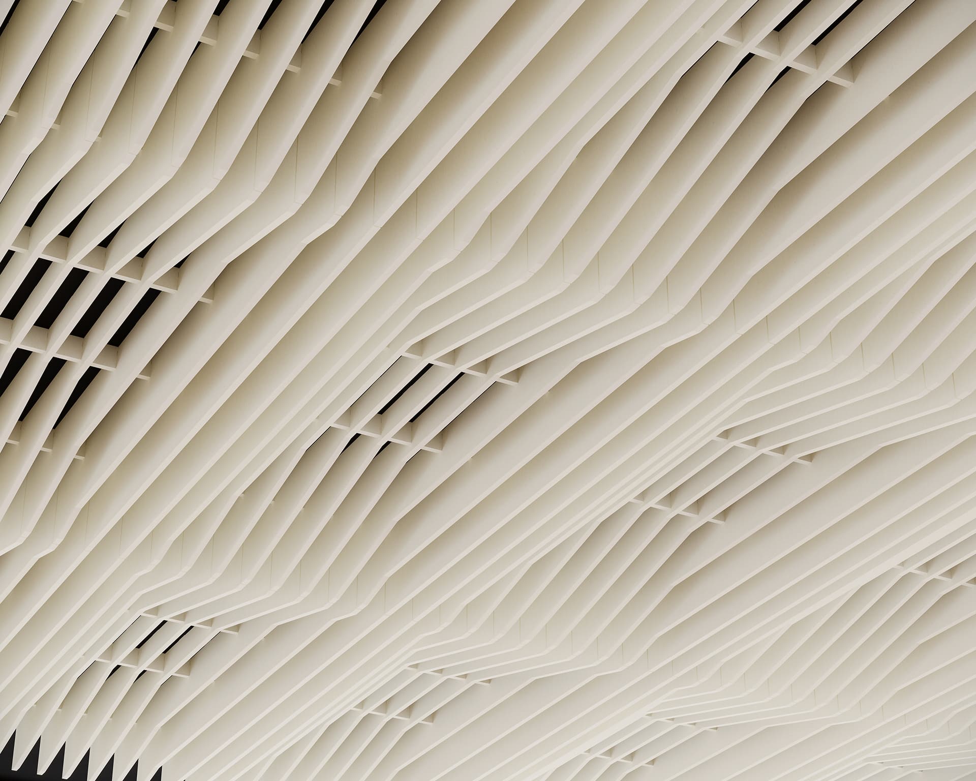 A close-up view of an acoustic felt ceiling with a geometric pattern featuring parallel, wavy lines and vertical slats. The design creates a visually striking texture and depth. The color scheme is a light beige or off-white.