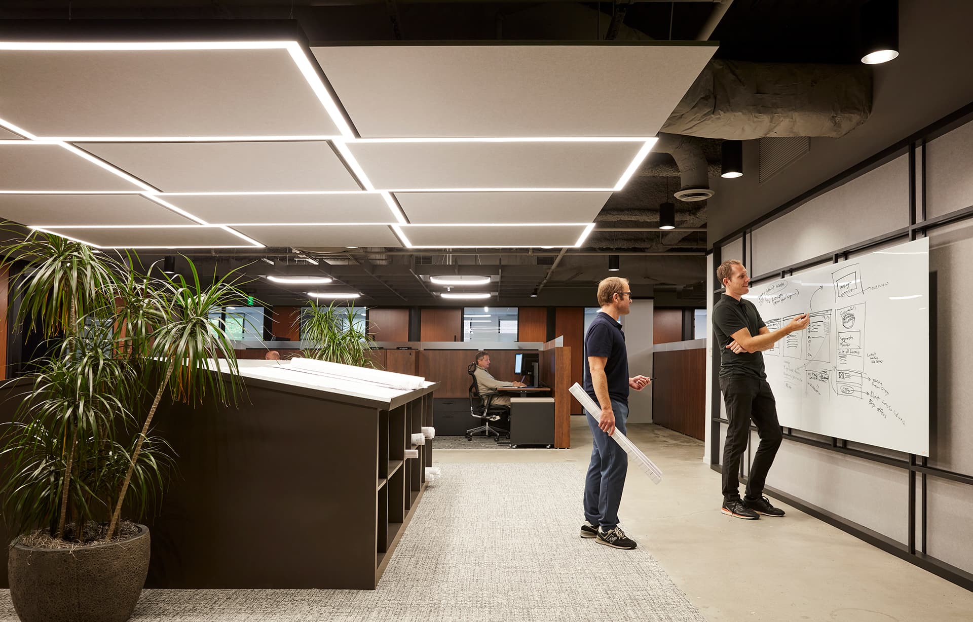 Two people are discussing at a whiteboard in a modern office, one writing while the other holds rolled papers. Nearby, another person works at a computer. There are large plants and modular ceiling lights around acoustic felt ceiling panels in the open-plan space.