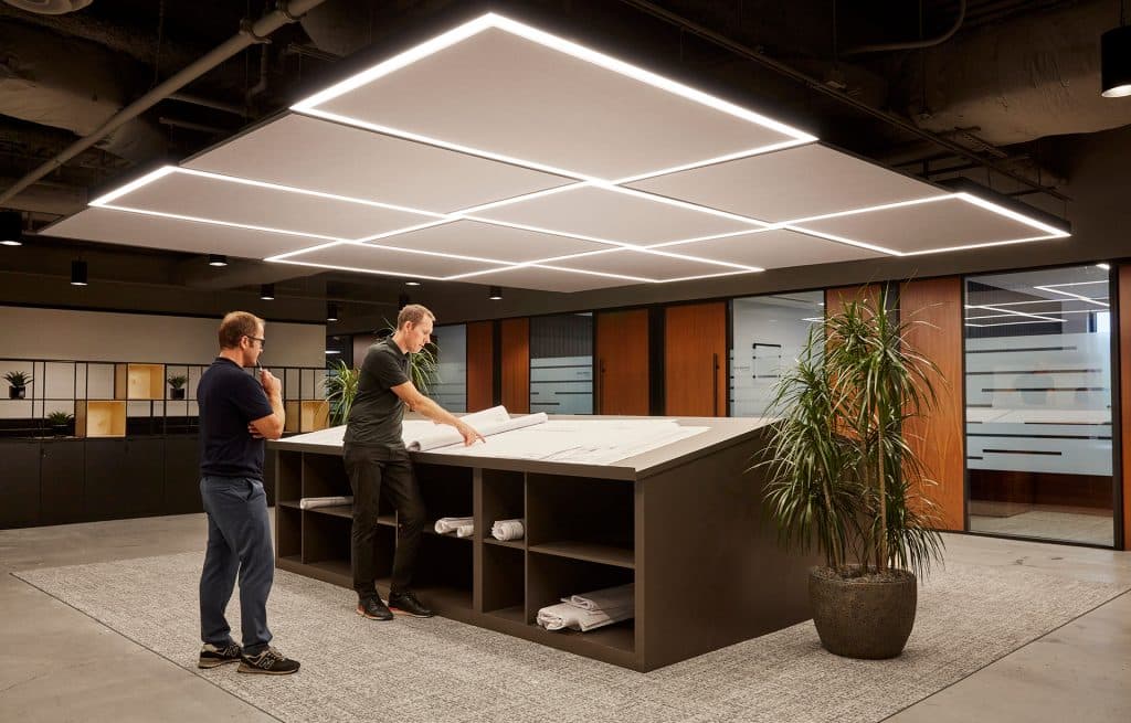 Two men stand at a large, slanted workspace reviewing architectural plans in a modern office. One wears a black shirt, and the other a blue shirt. The room is well-lit with geometric ceiling lights around acoustic felt ceiling panels and features large glass partitions and potted plants.