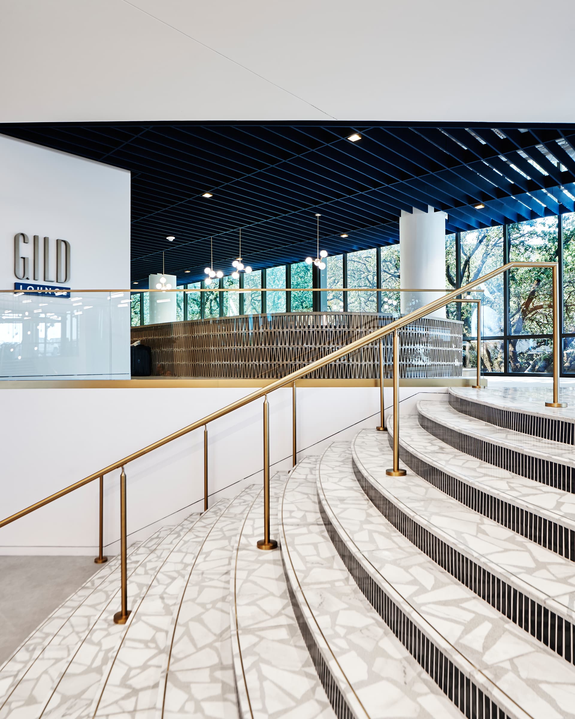 A modern interior features a curved staircase with a gold railing, mosaic-patterned steps, and a wall with the text "GILD." Large windows provide natural light, and the ceiling is dark blue acoustic felt with recessed lighting.