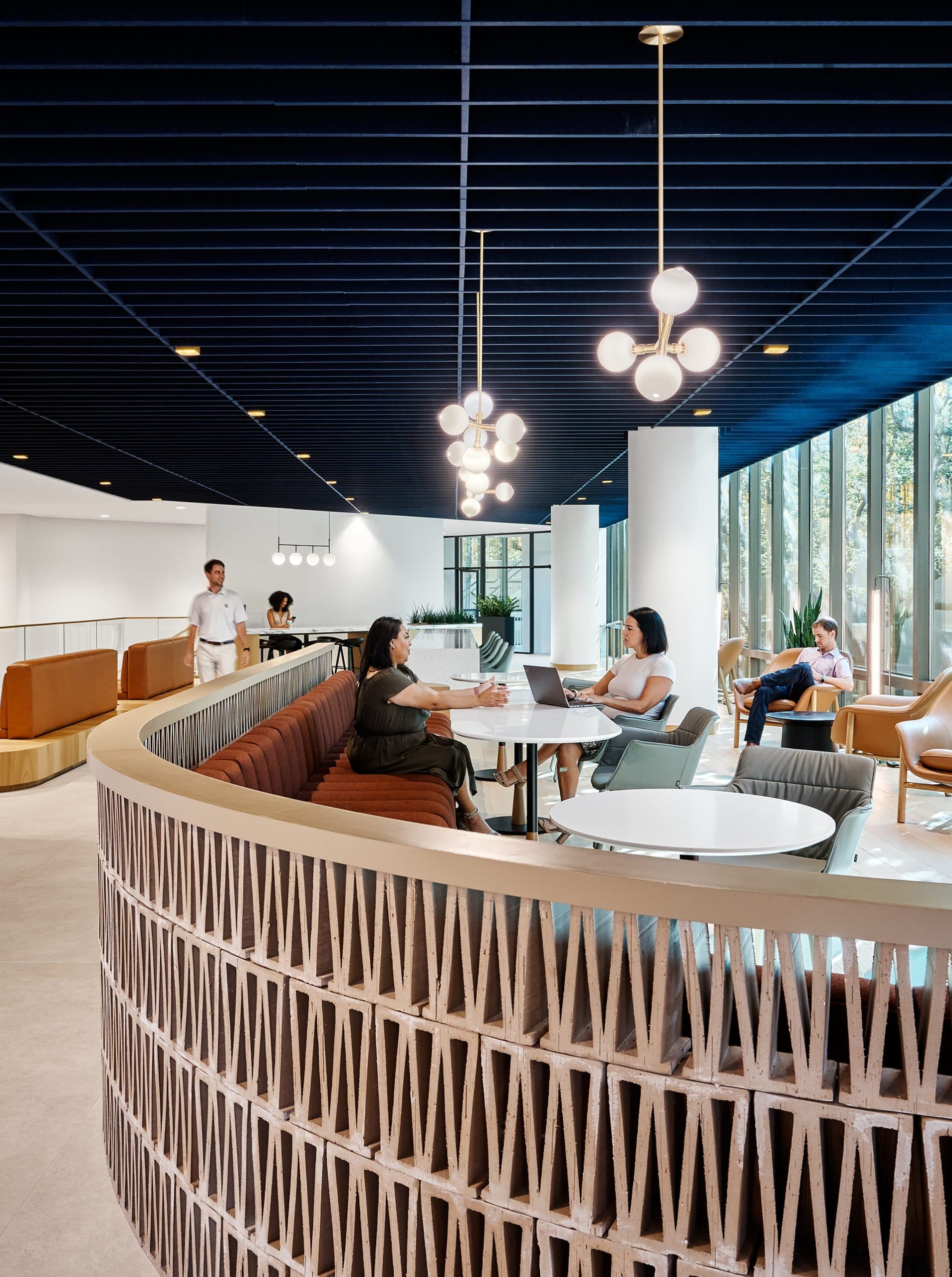 A modern office space with curved seating and a wooden partition. People are seated, engaged in conversation or on laptops. The ceiling is dark blue acoustic felt with spherical lights, and large windows provide natural light.