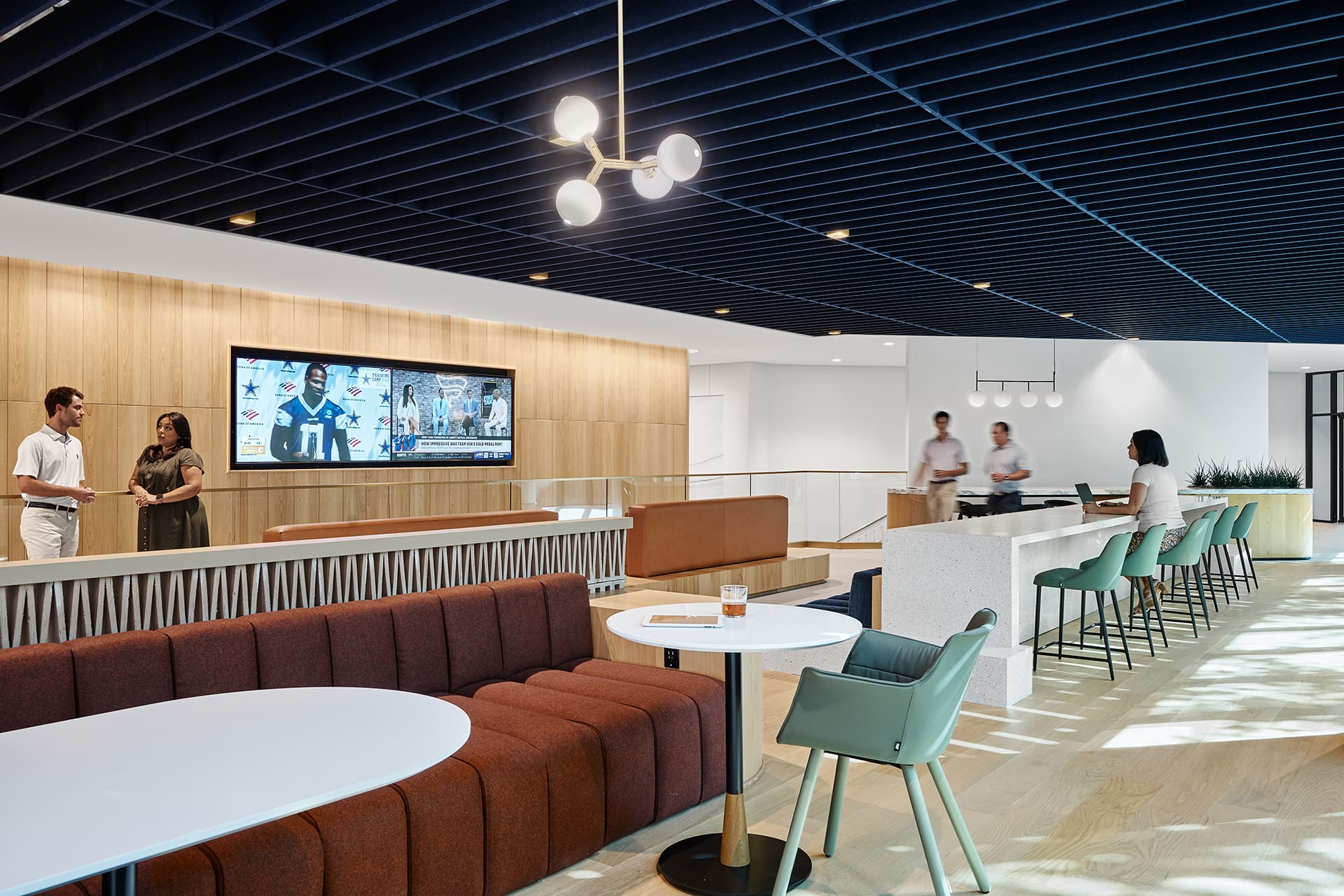 Modern office lounge with people conversing. Features include a brown sectional sofa, round tables, teal chairs, and a wall-mounted screen. A geometric light fixture hangs from a dark acoustic felt ceiling, and greenery is visible in the background.