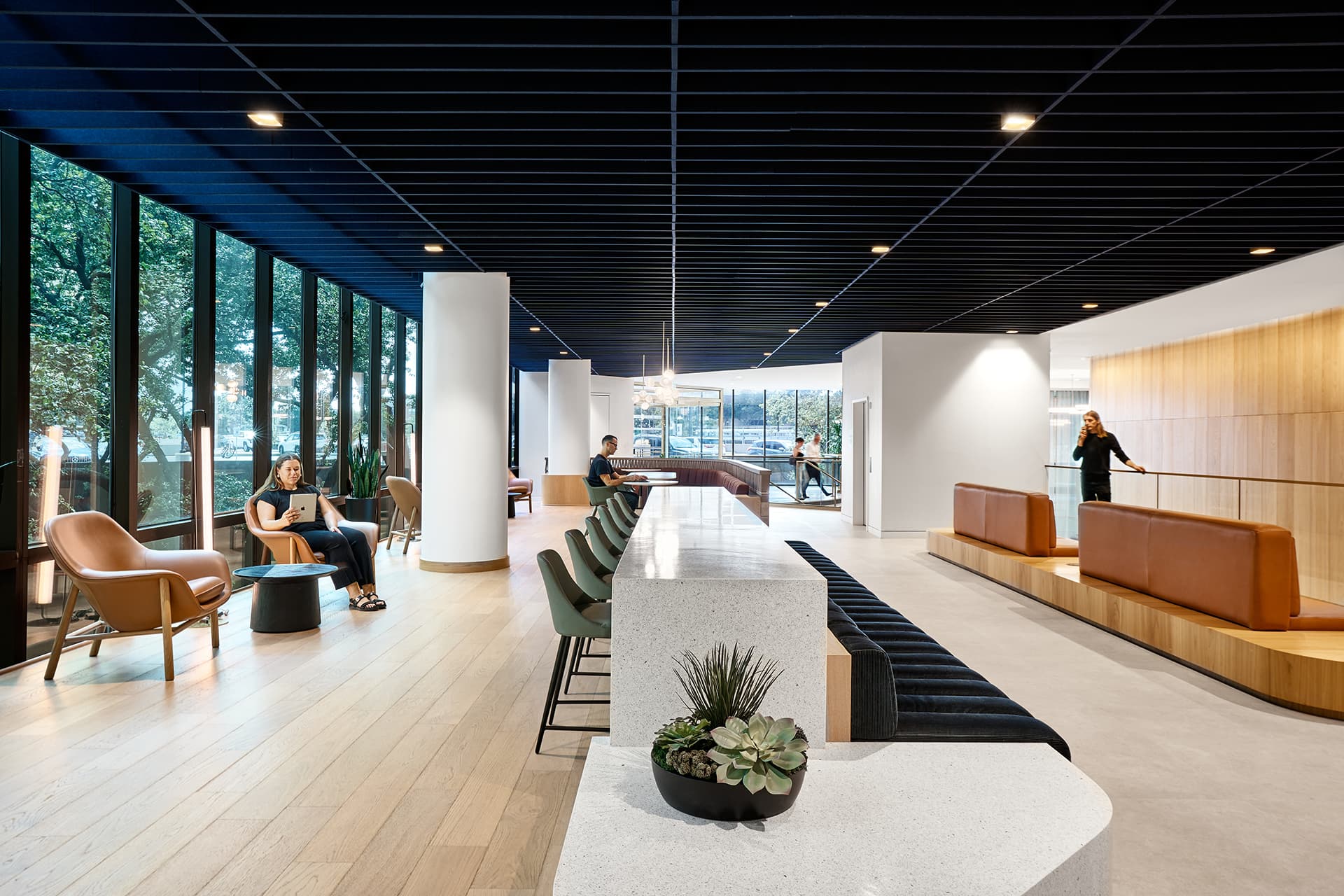 Modern office lounge with large windows and a dark blue acoustic felt ceiling. Features comfortable seating, a long white communal table, brown leather benches, and potted plants. People are sitting, and one person is standing in the background. Trees visible outside.